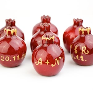 Könnte beinhalten: Sechs rote Keramik-Granatapfel-Ornamente mit goldenen Details. Jedes Ornament ist mit einem Datum und Initialen personalisiert.