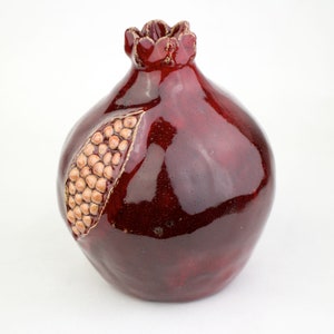 May include: A ceramic pomegranate sculpture in a deep burgundy glaze. The fruit features a detailed, textured surface with a section carved out to reveal the seeds. The top has a decorative, ruffled crown. A unique piece for home decor.