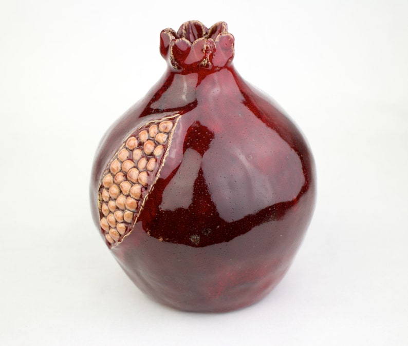 May include: A ceramic pomegranate sculpture in a deep burgundy glaze. The fruit features a detailed, textured surface with a section carved out to reveal the seeds. The top has a decorative, ruffled crown. A unique piece for home decor.