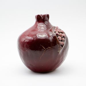 Puede incluir: Un jarr&oacute;n de cer&aacute;mica con forma de granada con un esmalte rojo oscuro y una superficie texturizada. El jarr&oacute;n tiene una peque&ntilde;a abertura en la parte superior y un dise&ntilde;o esculpido de semillas de granada en un lado.