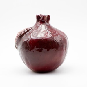 Puede incluir: Un jarr&oacute;n de cer&aacute;mica de color rojo oscuro con un acabado brillante. El jarr&oacute;n tiene forma de granada con un tallo detallado y una superficie ligeramente irregular.