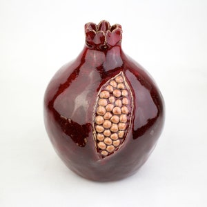May include: A ceramic pomegranate sculpture in a deep red glaze. The fruit features a detailed carving of the seeds, with a gold-colored border. The top has a crown-like design. This decorative piece is ideal for home decor.