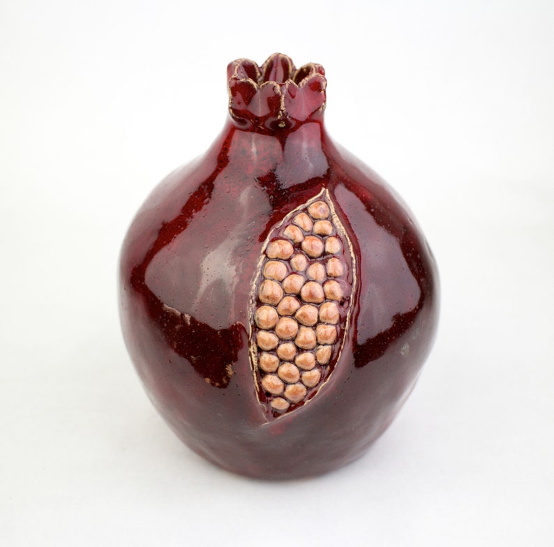 May include: A ceramic pomegranate sculpture in a deep red glaze. The fruit features a detailed carving of the seeds, with a gold-colored border. The top has a crown-like design. This decorative piece is ideal for home decor.