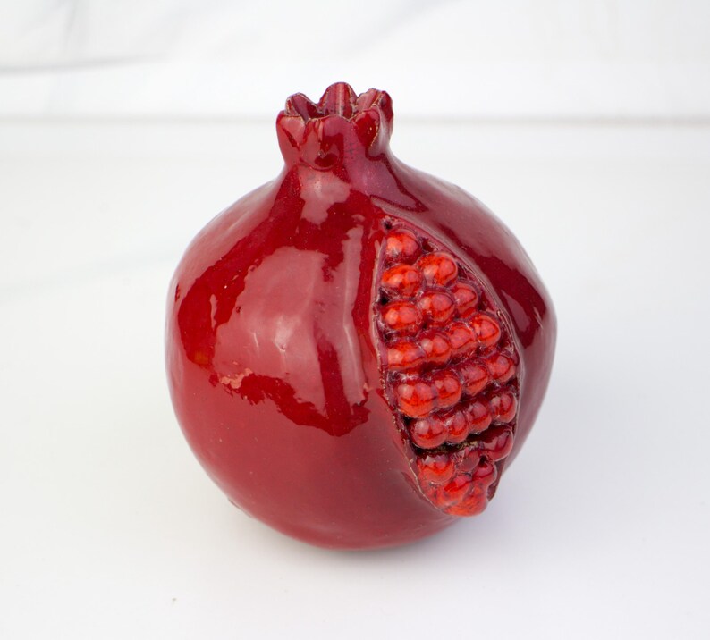 May include: A glossy, deep red pomegranate sculpture with a partially exposed interior revealing vibrant red seeds. The ceramic fruit has a textured surface and a detailed stem.