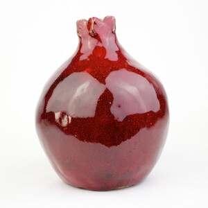 May include: A glossy, red ceramic vase with a pomegranate shape. The vase has a textured surface and a ruffled top. The deep red color and unique shape make it a decorative piece for any home.