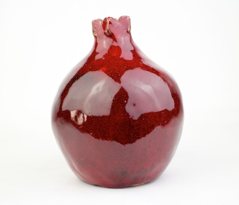 May include: A glossy, red ceramic vase with a pomegranate shape. The vase has a textured surface and a ruffled top. The deep red color and unique shape make it a decorative piece for any home.