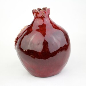 May include: A ceramic pomegranate-shaped vase with a glossy, deep red glaze. The vase has a textured surface and a detailed crown at the top. The design includes a textured, seed-like detail on the side.