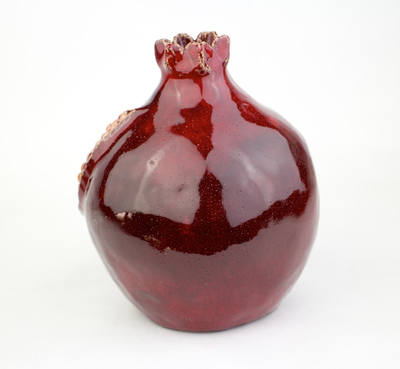 May include: A ceramic pomegranate-shaped vase with a glossy, deep red glaze. The vase has a textured surface and a detailed crown at the top. The design includes a textured, seed-like detail on the side.