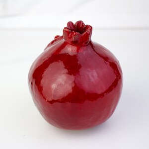 May include: A glossy, ceramic pomegranate sculpture in a deep red hue. The fruit features a textured crown and a smooth, reflective surface. This decorative piece is ideal for adding a touch of nature-inspired art to any space.