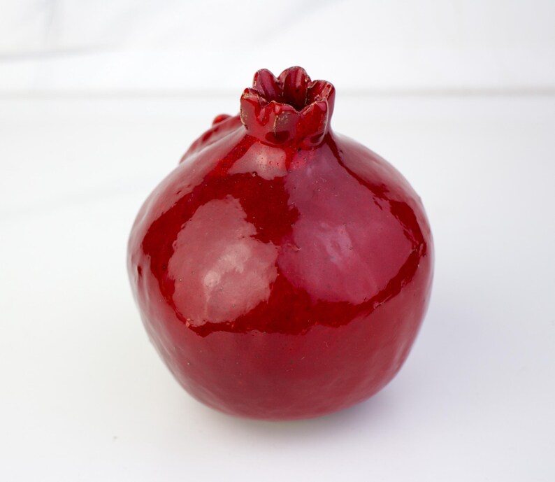 May include: A glossy, ceramic pomegranate sculpture in a deep red hue. The fruit features a textured crown and a smooth, reflective surface. This decorative piece is ideal for adding a touch of nature-inspired art to any space.