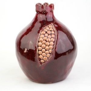 May include: A ceramic pomegranate sculpture in a deep burgundy glaze. The fruit features a detailed carving revealing the seeds, with a textured, light brown interior. The top has a decorative, scalloped edge.