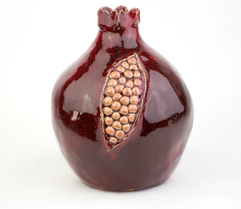 May include: A ceramic pomegranate sculpture in a deep burgundy glaze. The fruit features a detailed carving revealing the seeds, with a textured, light brown interior. The top has a decorative, scalloped edge.
