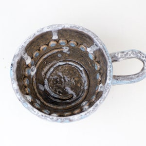 Op de afbeelding: Een keramische mok met een witte glazuur en bruine accenten. De mok heeft een uniek ontwerp met een spiraalvormig patroon en blauwe en witte stippen rond de rand.