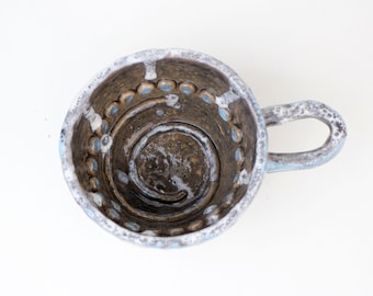 May include: A ceramic mug with a white glaze and brown accents. The mug has a unique design with a spiral pattern and blue and white dots around the rim.