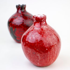 May include: Two red ceramic vases shaped like pomegranates. One vase has a black and red glaze, while the other has a red glaze with white speckles.