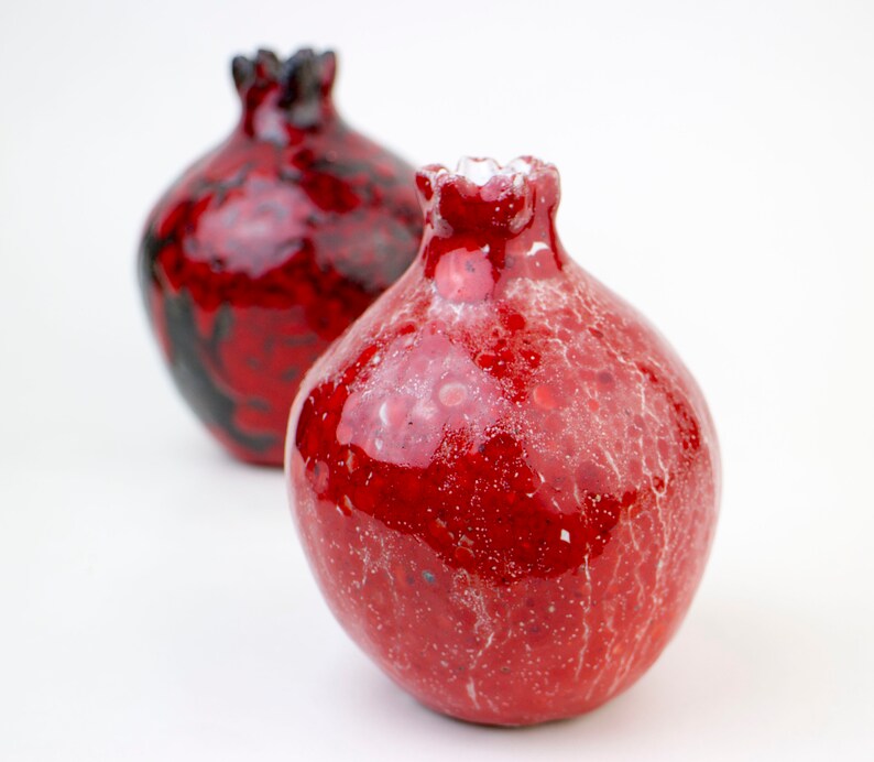 May include: Two red ceramic vases shaped like pomegranates. One vase has a black and red glaze, while the other has a red glaze with white speckles.