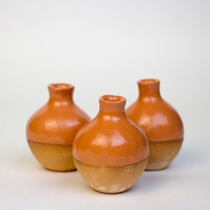 May include: Three small, round ceramic vases with a two-tone design. The top half of each vase is a glossy orange, while the bottom half is a matte tan. The vases have a narrow neck and a rounded body, and are arranged against a white background.