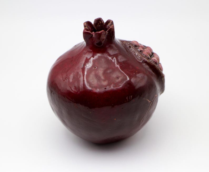Puede incluir: Un jarr&oacute;n de cer&aacute;mica con forma de granada de color rojo oscuro con un acabado brillante. El jarr&oacute;n tiene un dise&ntilde;o texturizado detallado que se asemeja a la piel de una granada.