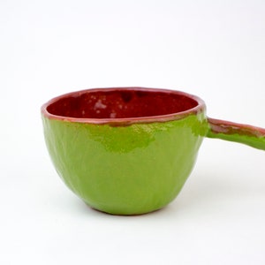 Puede incluir: Un pequeño cuenco de cerámica verde con un borde rojo y una asa. El cuenco tiene una superficie texturizada.
