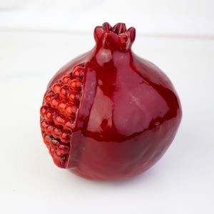 May include: A glossy, deep red pomegranate sculpture with a partially exposed interior revealing numerous small, red seeds. The fruit's exterior has a smooth, reflective surface, and the top features a crown-like structure.