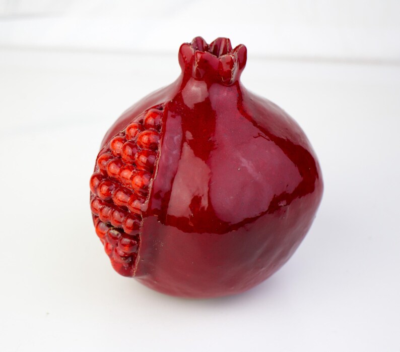 May include: A glossy, deep red pomegranate sculpture with a partially exposed interior revealing numerous small, red seeds. The fruit's exterior has a smooth, reflective surface, and the top features a crown-like structure.
