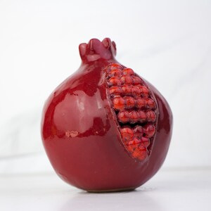 May include: A ceramic pomegranate sculpture in a deep red glaze. The fruit is partially open, revealing a cluster of red seeds. The sculpture has a glossy finish and sits on a white surface.