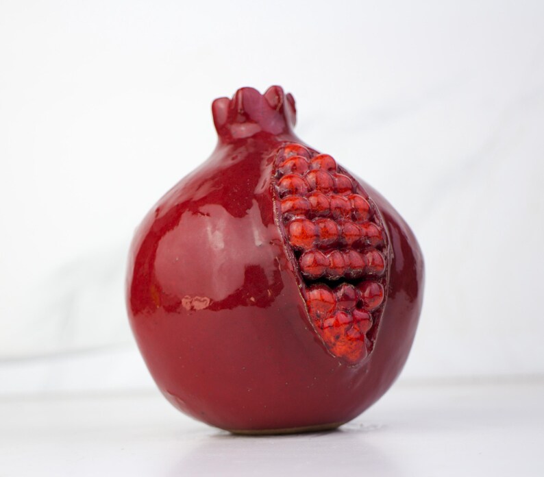 May include: A ceramic pomegranate sculpture in a deep red glaze. The fruit is partially open, revealing a cluster of red seeds. The sculpture has a glossy finish and sits on a white surface.