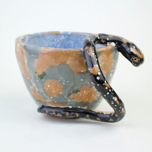 以下が含まれることがあります： ユニークな蛇の形をした取っ手が付いた手作りのセラミックマグ。マグカップは、茶色、グレー、青の色合いのまだら模様のデザインです。蛇の取っ手は黒色で、白と茶色の斑点があり、遊び心のあるデザインです。