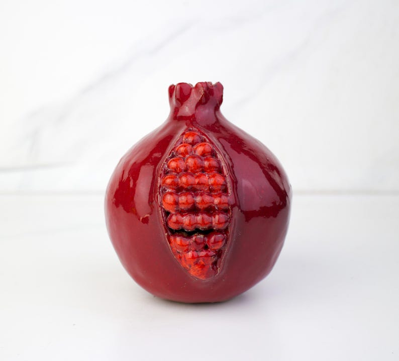 May include: A glossy, red pomegranate sculpture with a detailed, open center revealing the fruit's seeds. The sculpture has a smooth, reflective surface and is set against a white background. The pomegranate is a symbol of abundance and fertility.