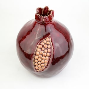 May include: A ceramic pomegranate sculpture in a deep red glaze. The fruit features a detailed section revealing the seeds, with gold-colored accents. The top has a crown-like design. This decorative piece is ideal for home decor.