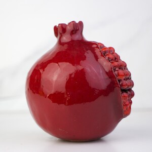 May include: A glossy, red ceramic pomegranate sculpture. The fruit's exterior is a deep, rich red with a shiny glaze. One side reveals a textured, seed-like detail. The pomegranate has a rounded shape with a textured crown.