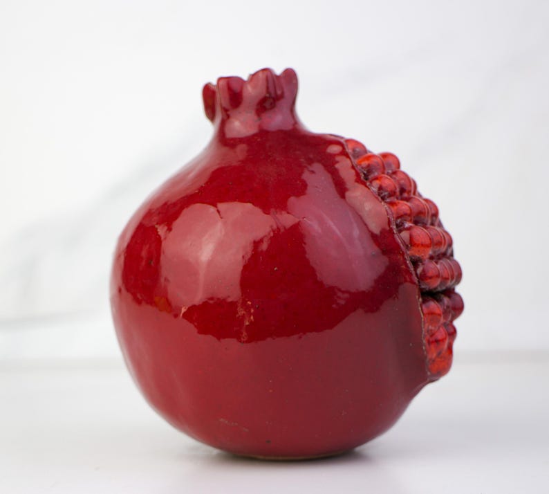 May include: A glossy, red ceramic pomegranate sculpture. The fruit's exterior is a deep, rich red with a shiny glaze. One side reveals a textured, seed-like detail. The pomegranate has a rounded shape with a textured crown.