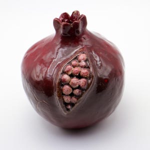 Puede incluir: Una figurita de cer&aacute;mica en forma de granada con un esmalte rojo oscuro y una representaci&oacute;n detallada de las semillas en su interior. La figurita es un objeto decorativo.