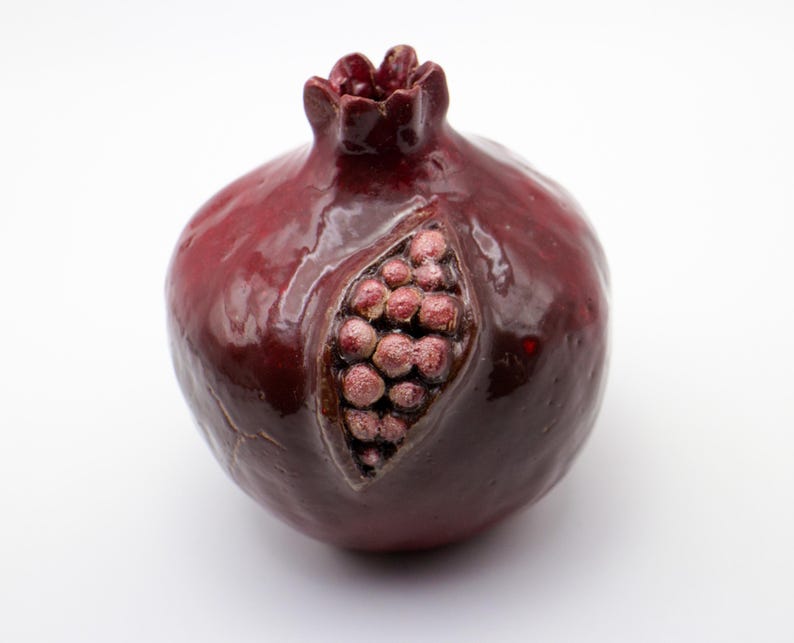 Puede incluir: Una figurita de cer&aacute;mica en forma de granada con un esmalte rojo oscuro y una representaci&oacute;n detallada de las semillas en su interior. La figurita es un objeto decorativo.
