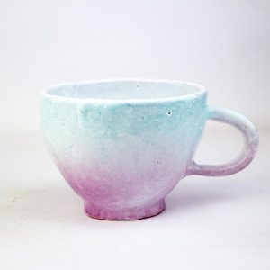 May include: A small, white ceramic mug with a light blue and purple ombre glaze. The mug has a rounded shape and a handle.
