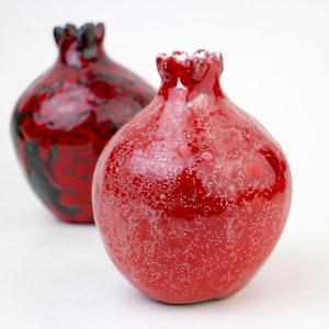 May include: Two red ceramic pomegranate-shaped vases. One vase has a black and red glaze, while the other has a red glaze with white speckles.