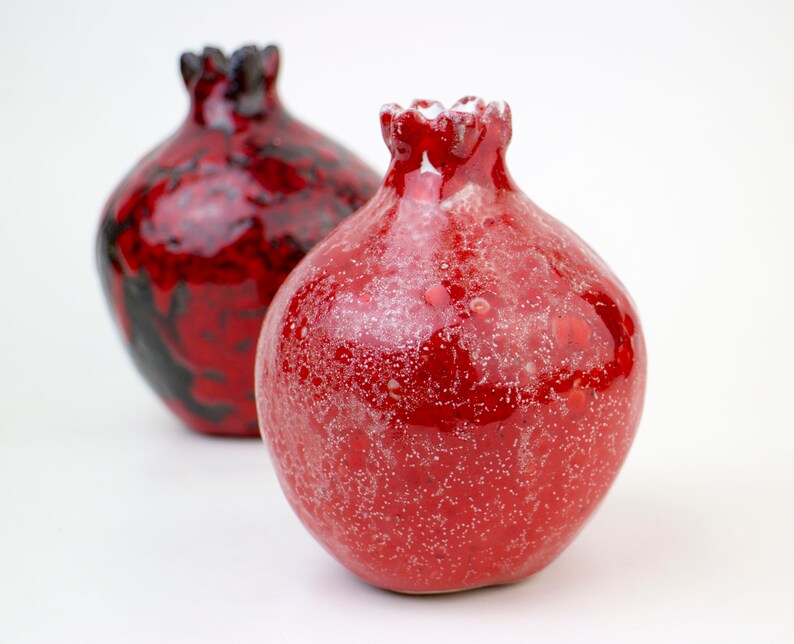 May include: Two red ceramic pomegranate-shaped vases. One vase has a black and red glaze, while the other has a red glaze with white speckles.