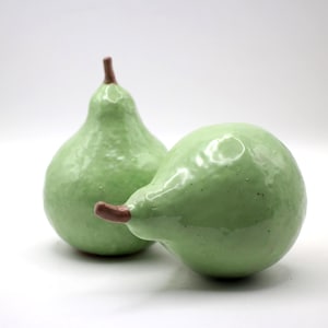 Puede incluir: Dos peras de cerámica verde claro con tallos marrones. Una pera está en posición vertical y la otra está acostada de lado. Las peras tienen un acabado brillante sobre un fondo blanco.