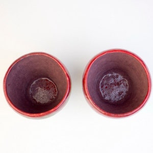 May include: Two red ceramic cups with a dark purple glaze inside. The cups are empty and sitting on a white surface.