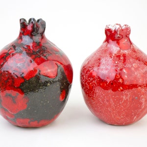 May include: Two red ceramic pomegranate-shaped vases. One vase has a black glaze with red accents, while the other vase is a solid red with white speckles.