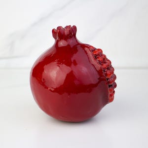 May include: A glossy, deep red pomegranate sculpture. The fruit features a textured, seed-filled section on its side and a crown-like top. The pomegranate is a decorative item, likely for home decor.