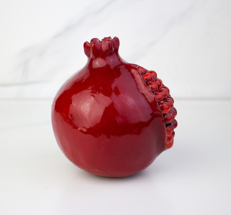 May include: A glossy, deep red pomegranate sculpture. The fruit features a textured, seed-filled section on its side and a crown-like top. The pomegranate is a decorative item, likely for home decor.