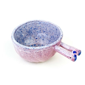 Puede incluir: Un pequeño cuenco de cerámica rosa y blanco con un esmalte moteado. El cuenco tiene dos asas azules con forma de pequeñas orejas redondeadas.