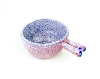 May include: A small, pink and white ceramic bowl with a speckled glaze. The bowl has two blue handles that are shaped like small, rounded ears.