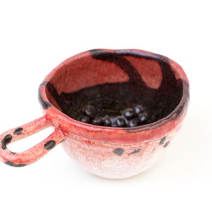 Puede incluir: Un pequeño cuenco de cerámica rojo y negro con asa. El cuenco tiene un esmalte moteado y un interior marrón oscuro.