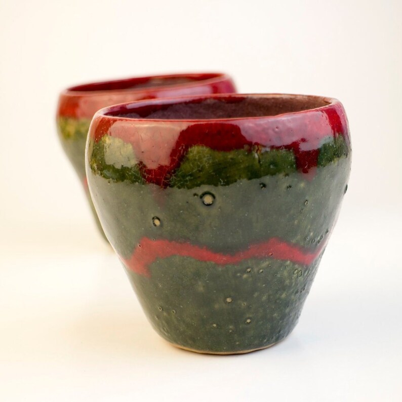 May include: Two handmade ceramic mugs with a green and red glaze. The mugs have a unique, speckled finish and are perfect for adding a touch of rustic charm to your kitchen.