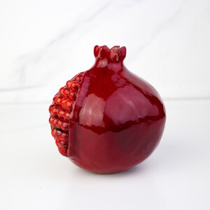 May include: A glossy, deep red pomegranate sculpture with a textured, seed-filled section on one side. The fruit-shaped object has a detailed crown and a smooth, reflective surface. The pomegranate is a decorative item.