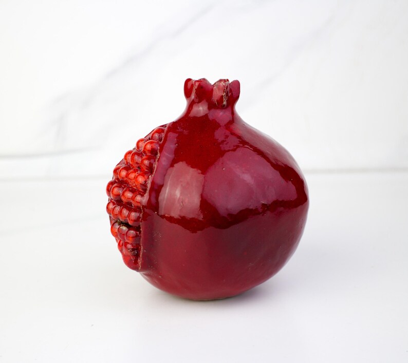 May include: A glossy, deep red pomegranate sculpture with a textured, seed-filled section on one side. The fruit-shaped object has a detailed crown and a smooth, reflective surface. The pomegranate is a decorative item.