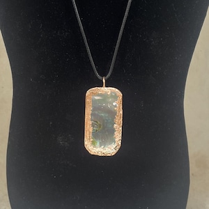 May include: A rectangular pendant necklace with a copper-colored frame and a clear, iridescent center. The pendant hangs from a black cord. The necklace is displayed on a black mannequin.