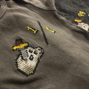 May include: A dark gray t-shirt featuring a cross-stitched design of a pixelated skull wearing a yellow hat, accompanied by the embroidered text 1/1. A second black garment is partially visible in the background with the embroidered text 99/99.